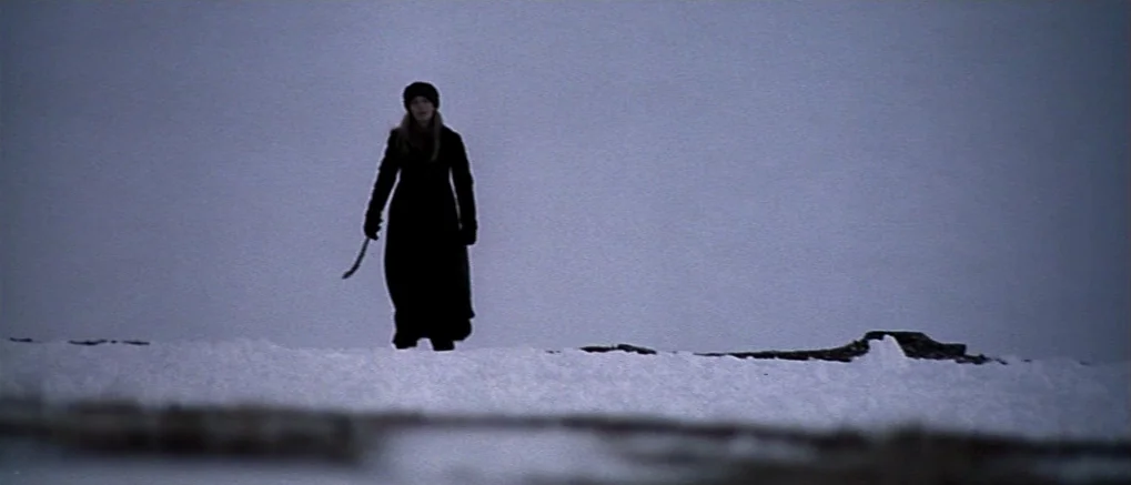 The Claim (2000) movie still: fullbody — A vast, flat snowy landscape stretches under a thick, overcast grey sky. A woman dressed in dark winter…