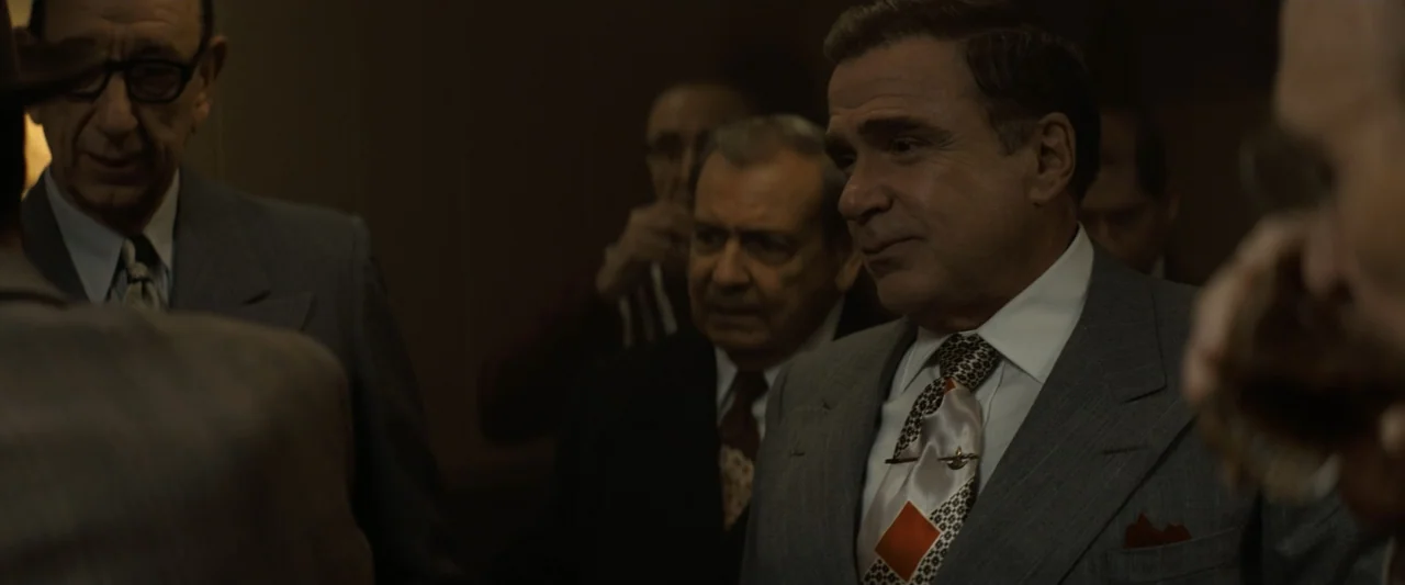 The Alto Knights (2025) movie still: medium — A dimly lit interior room with dark wood paneling backdrop. Three men in 1950s business attire stand…