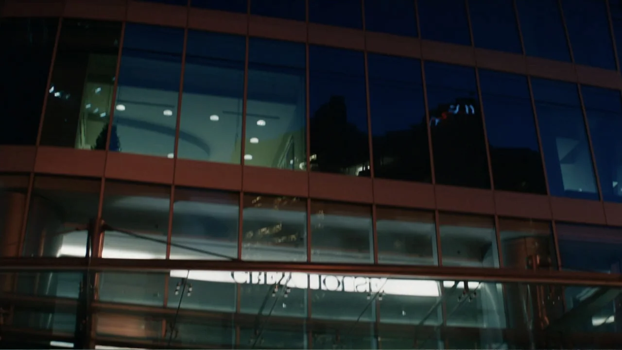Fifty Shades of Grey (2015) movie still: establishing — A modern glass-fronted office building at night with lit interior workspaces visible.…