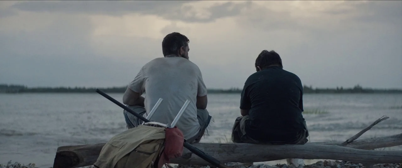 The Peanut Butter Falcon (2019) movie still: fullbody — Two men sit together on a weathered log by a large, calm lake. A canvas backpack and a…