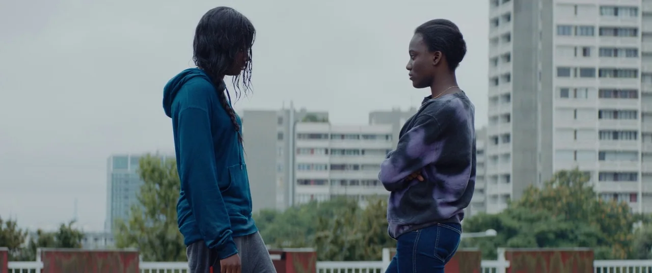 Girlhood (2014) movie still: medium — An urban rooftop terrace overlooks tall concrete apartment blocks under an overcast grey sky. Two young women…