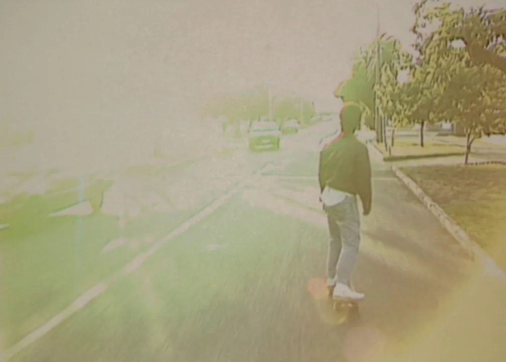 No (2012) movie still: fullbody — A person rides a skateboard along a paved path next to a residential road. Several trees stand on the right side…