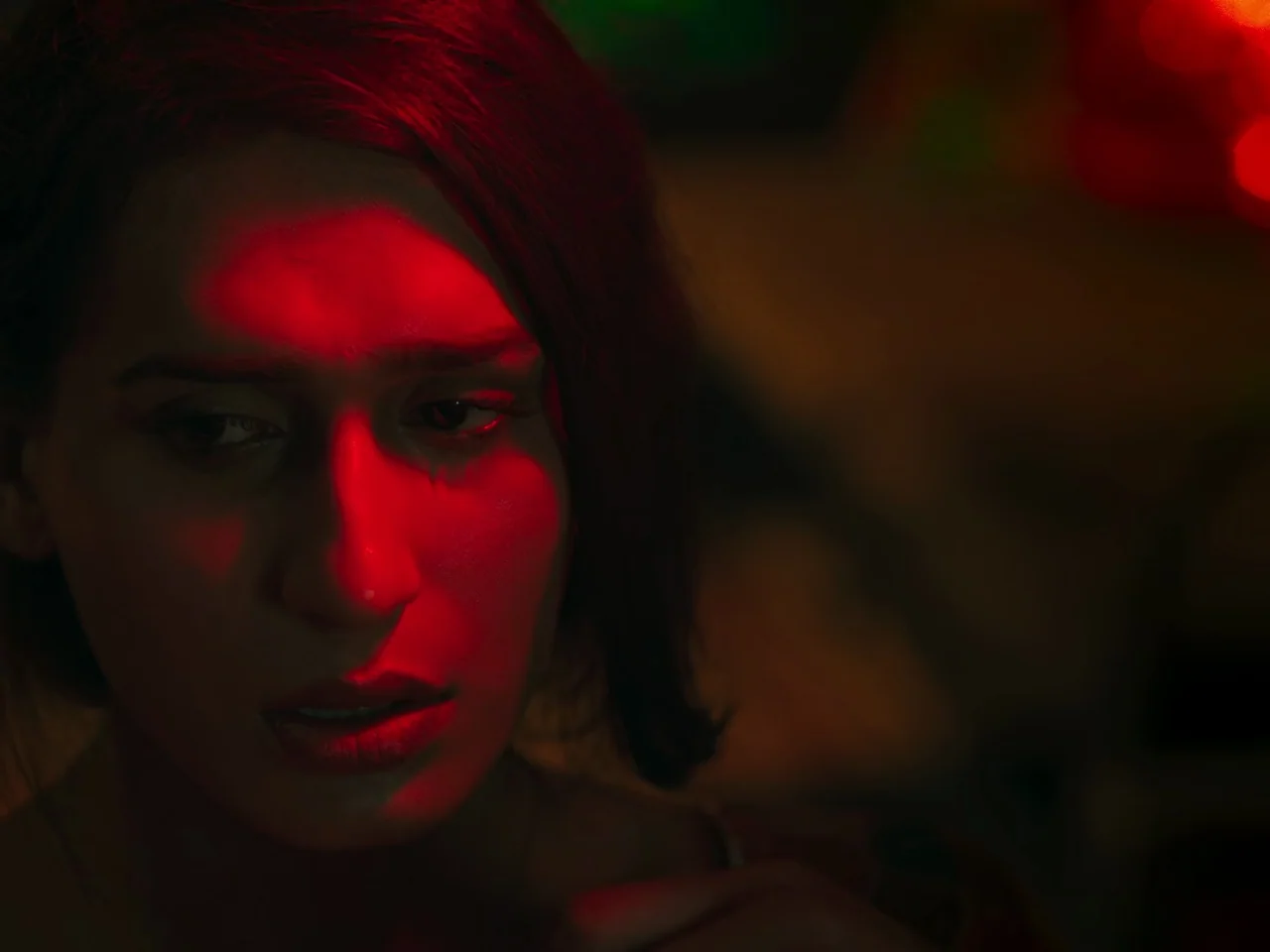 Joyland (2022) movie still: closeup — A dark indoor space with minimal ambient light and deep shadows throughout the frame. A young woman with dark…