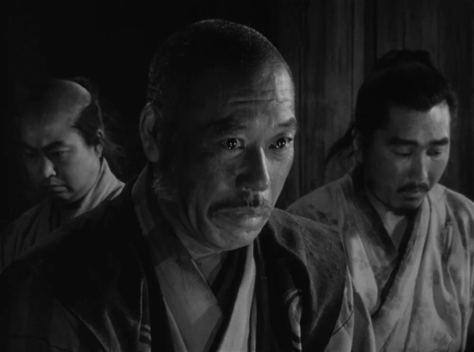 Seven Samurai (1954) movie still: medium — An interior space with wooden walls in a dimly lit, austere setting. Three men in traditional Japanese…