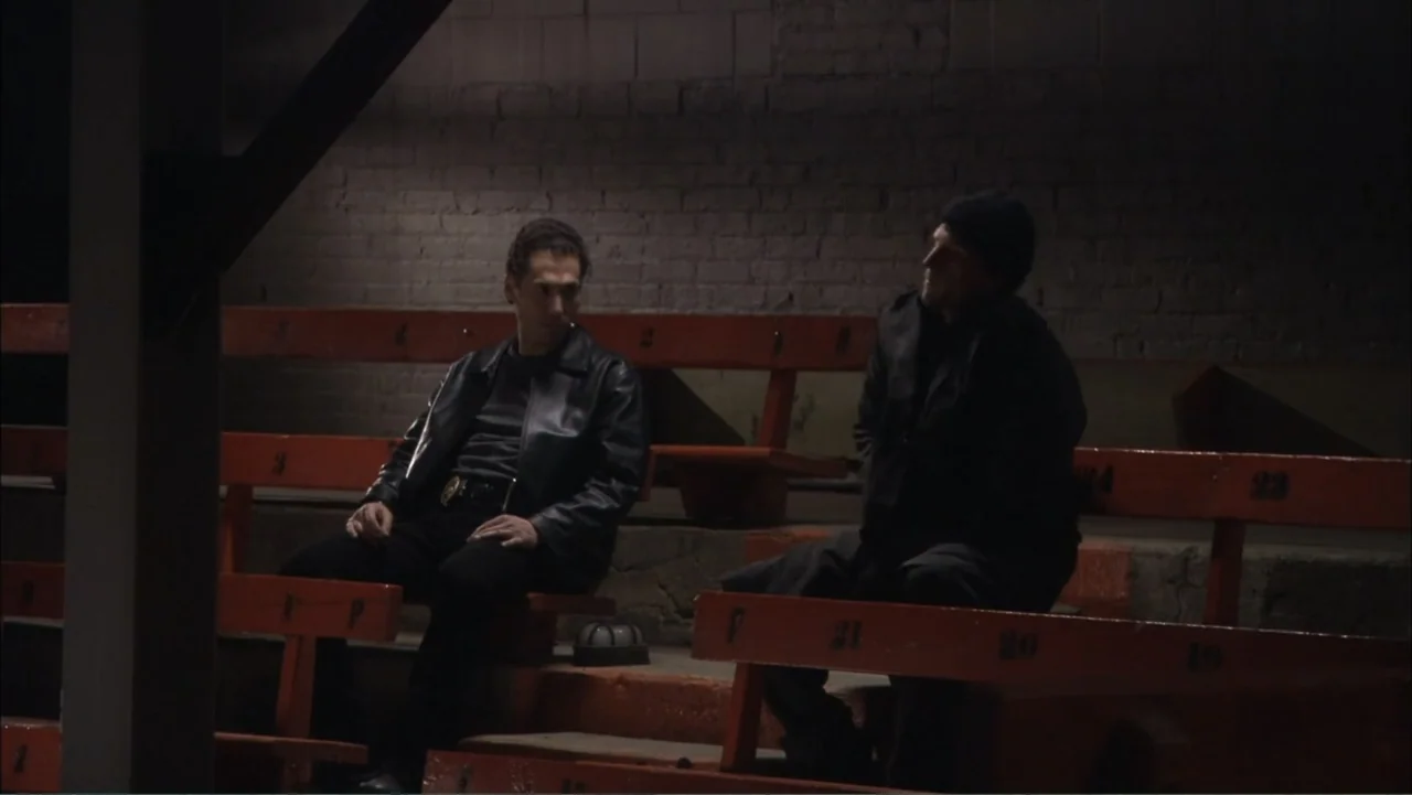 O código oculto (2025) movie still: medium — Two men sit together on long orange wooden bleachers in a dimly lit space. The wall behind them is…