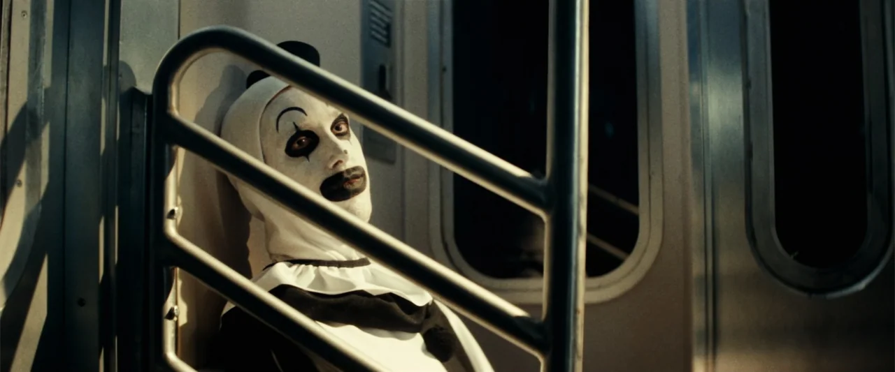Terrifier 3 (2024) movie still: medium — A dark subway car with metallic walls and a steel handrail in the foreground. A person dressed in white…
