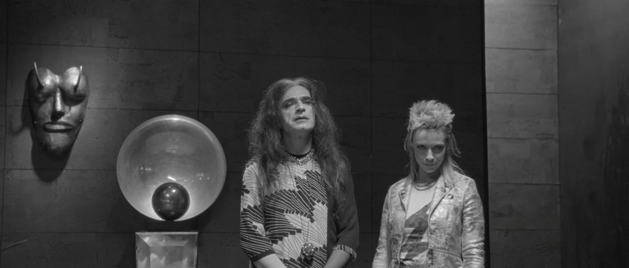 She Is Conann (2023) movie still: medium — A dark, modern interior features a tiled wall decorated with a metal horned mask. Two people stand in the…