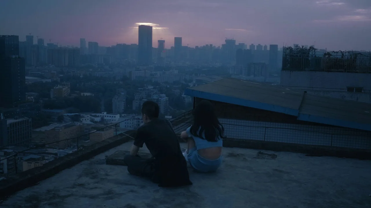 The Last Year of Darkness (2023) movie still: wide — A flat rooftop overlooks a vast, densely packed cityscape during the late evening hour. A young…