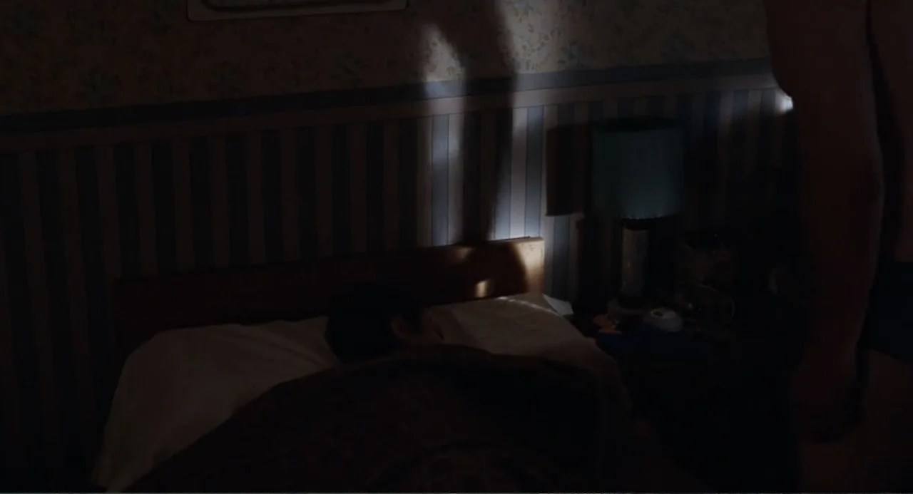 La Ciénaga (2001) movie still: medium — A dim bedroom with patterned floral wallpaper and a small lamp by the bed. One person stands partially…