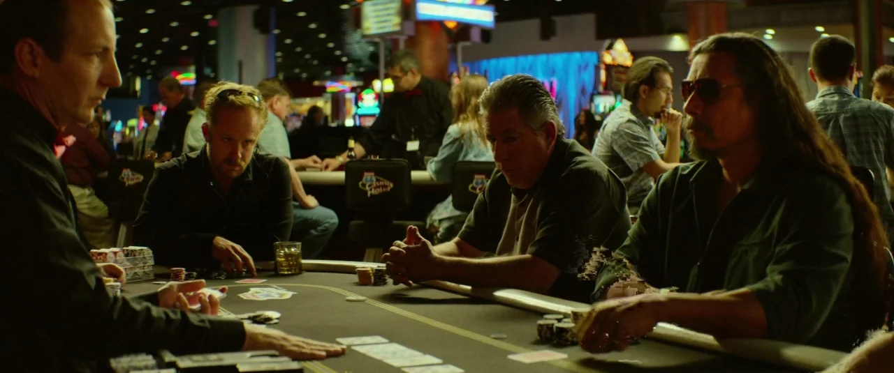 Hell or High Water (2016) movie still: medium — A group of men play poker in a dark, neon-lit casino at night. They sit at a long table with chips…