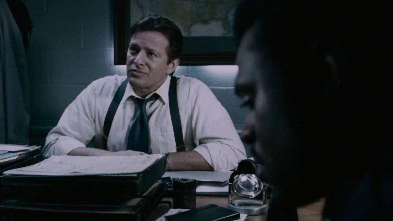Saw IV movie still: medium — A dimly lit room features a man sitting at a desk behind a file folder. The man is positioned in the center and looks…