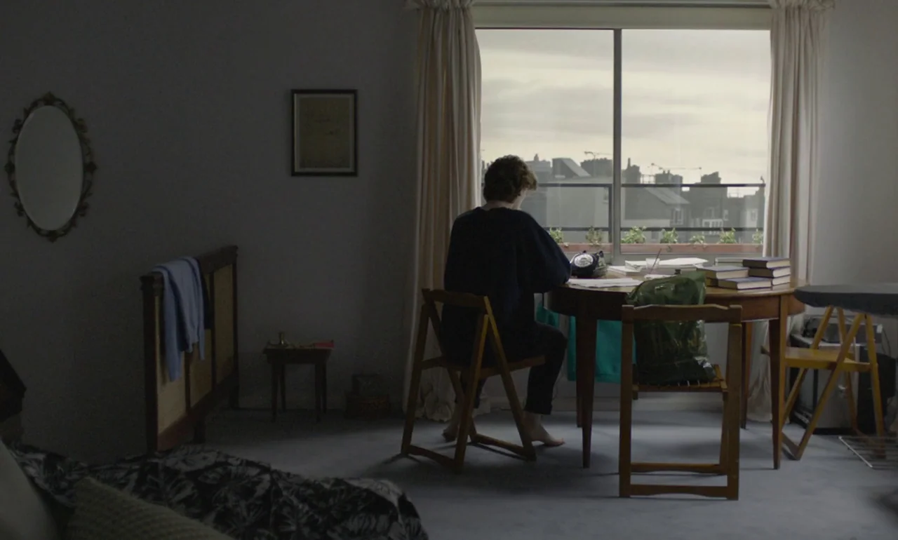 The Souvenir (2019) movie still: medium — An apartment interior features a person sitting at a wooden table near a window. Sunlight filters through…