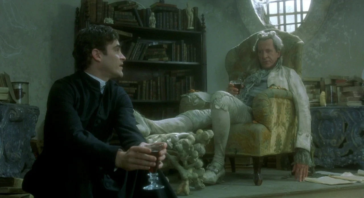 Quills (2000) movie still: medium — A cluttered, dim study contains dark wooden bookshelves filled with aged, leather-bound volumes. A younger man…