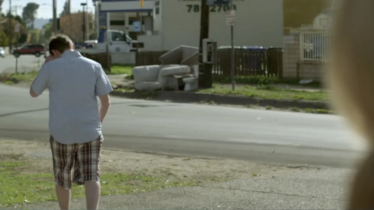 Free Samples (2013) movie still: medium — A suburban street scene with a paved road and a discarded couch on the side. A man stands in the left…