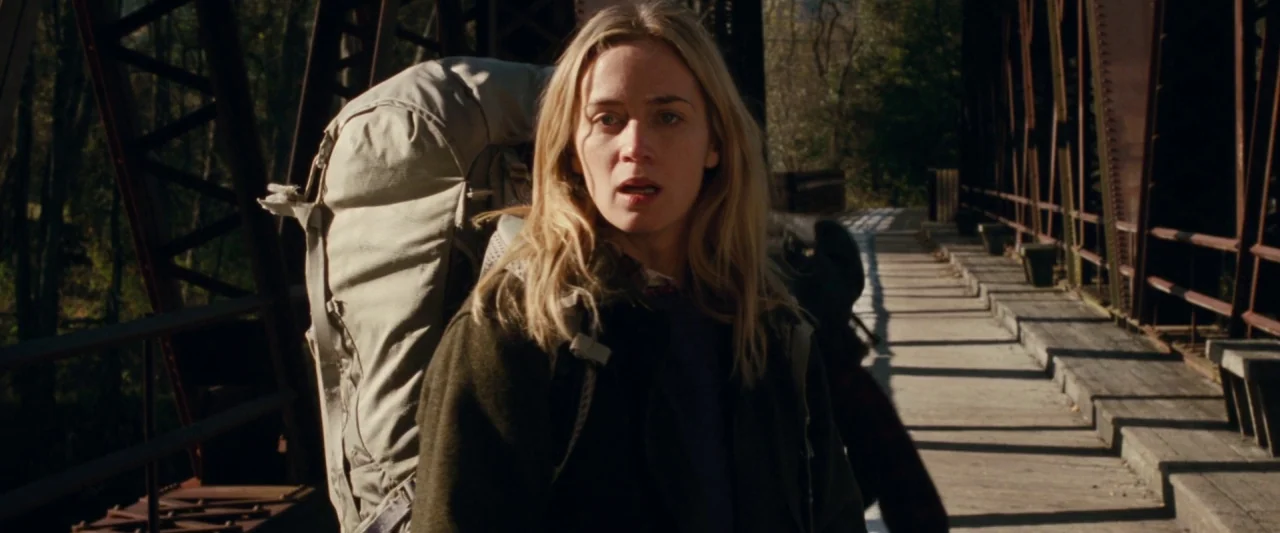 A Quiet Place (2018) movie still: medium — A woman walks across a metal truss bridge outdoors in daylight. She wears a large hiking backpack and…