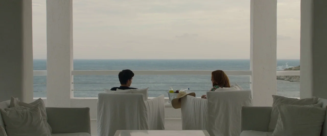 Greed (2019) movie still: wide — A white-pillared terrace overlooks a calm blue sea under an overcast sky. A man and a woman sit in white-covered…