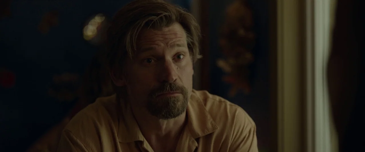 Small Crimes (2017) movie still: closeup — A dimly lit room with dark blue walls serves as the backdrop. A man in a tan shirt looks toward an unseen…
