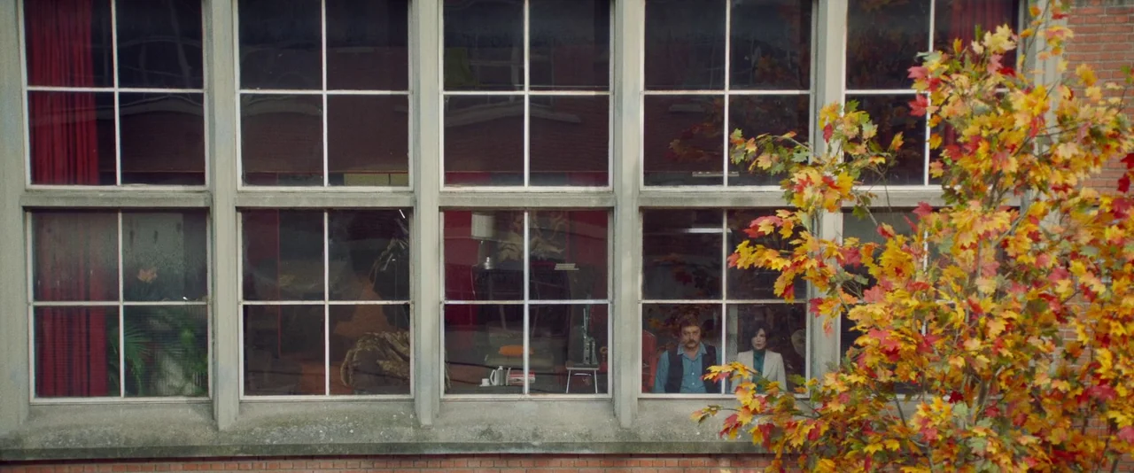 Peter Von Kant (2022) movie still: wide — A brick building facade features a large window with multiple panes and red curtains. A man and a woman…