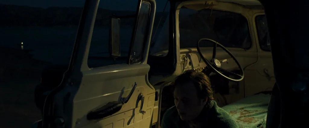 Zodiac (2007) movie still: medium — An interior of an older truck parked by a dark body of water at night. A young man sits in the driver seat with…