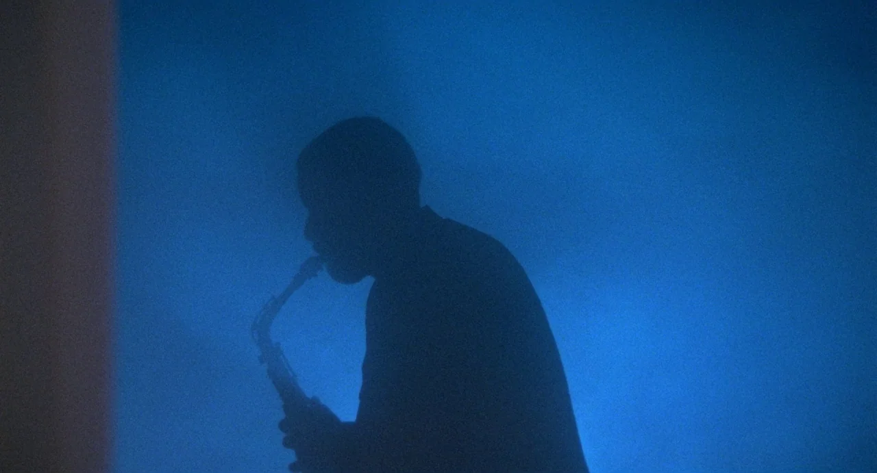 Def By Temptation (1990) movie still: medium — A dimly lit, hazy interior space is dominated by a deep blue light source. A musician stands in…