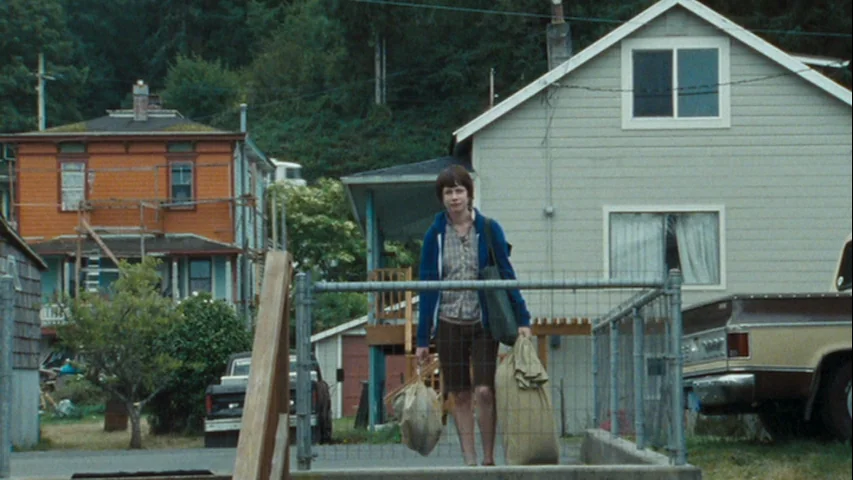 Wendy and Lucy (2008) movie still: fullbody — A residential neighborhood features houses, parked vehicles, and a chain-link fence. A woman walks…
