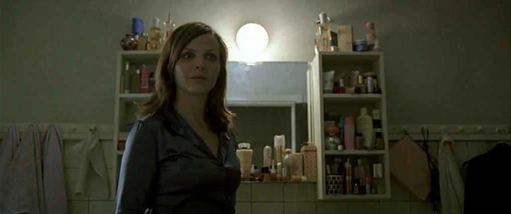 Next Door (2005) movie still: medium — A utilitarian bathroom features tiled walls and open shelving cluttered with various cosmetic bottles. A…