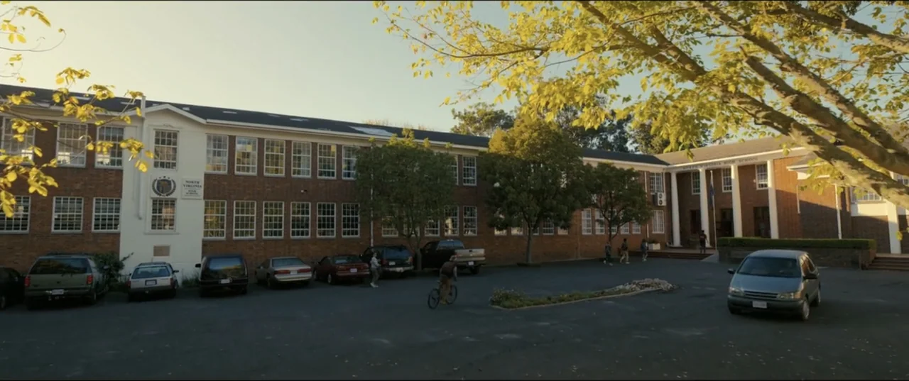 My Spy The Eternal City (2024) movie still: wide — A two-story brick high school building sits behind an asphalt parking lot with multiple vehicles.…
