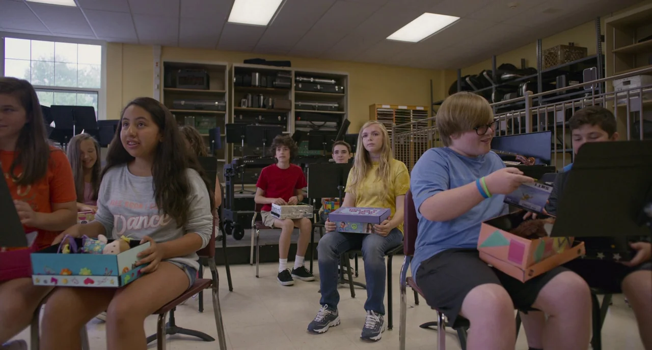 Eighth Grade (2018) movie still: medium — A school music room with tiered risers, musical instrument cases, and metal shelving units. A group of…
