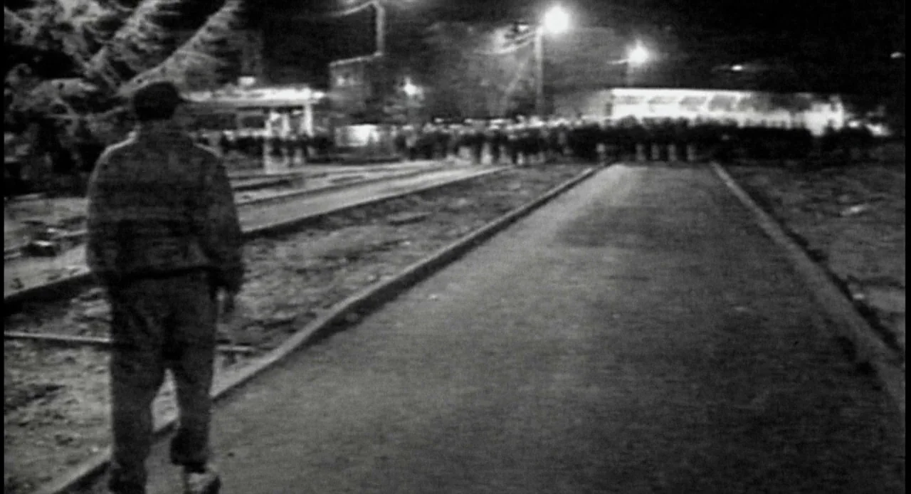 La Haine (1995) movie still: fullbody — A man walks alone along dark tram tracks at night under harsh artificial lighting. A blurry crowd of people…