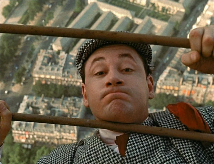 Zazie Dans Le Métro (1960) movie still: closeup — An aerial view of an urban landscape is visible behind metal bars. A man in a patterned suit looks…