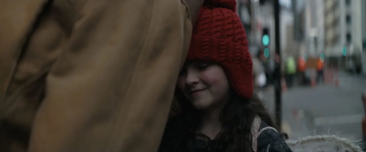 Wolf Man (2025) movie still: closeup — A city street at night with a blurred background of traffic lights and buildings. A young girl with a red…