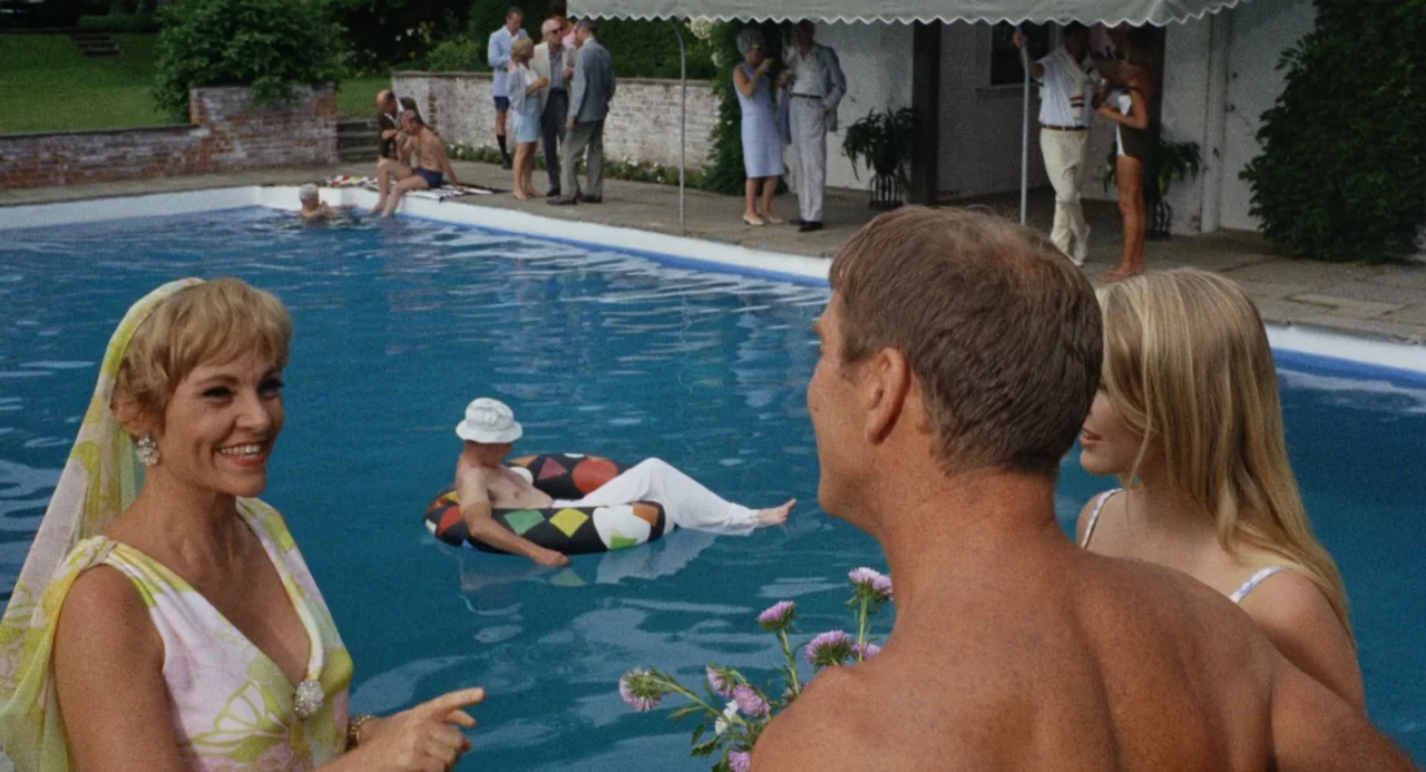 The Swimmer (1968) movie still: medium — A sunny poolside social gathering features multiple people standing on a paved patio. An older man in a…