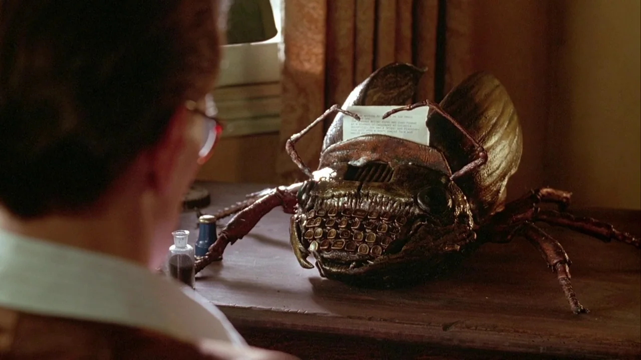 Naked Lunch (1991) movie still: medium — A dimly lit room contains a wooden desk with a large insect-shaped typewriter. A man sits facing the desk,…