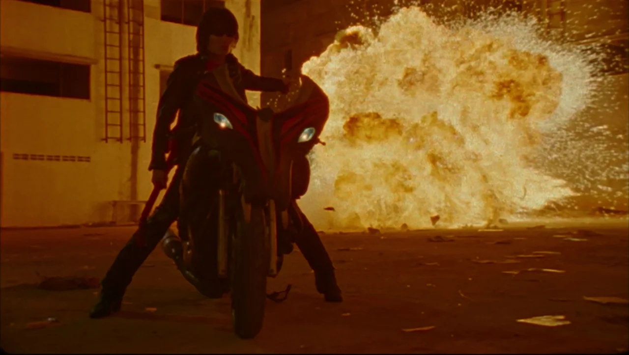 Wild Zero (1999) movie still: fullbody — A dark, paved industrial area serves as the site for a large fiery explosion. A person in black clothing…