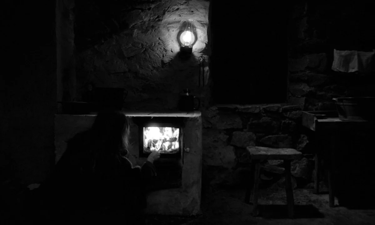 The Turin Horse (2011) movie still: medium — A dark, rustic room with thick stone walls and a small wood-burning stove. A woman kneels in front of…