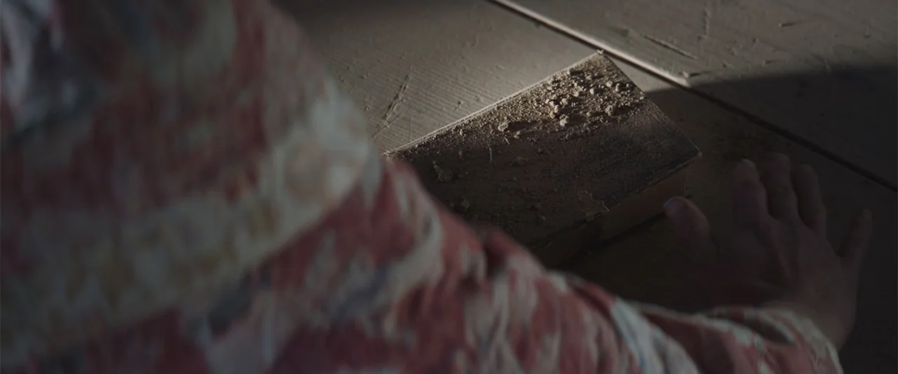 The Wind (2005) movie still: closeup — A dusty book lies on a wooden floor illuminated by a shaft of light. A person reaches toward the book while…