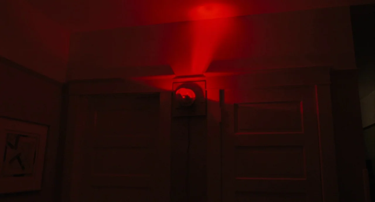 When You Finish Saving The World (2022) movie still: wide — A dim interior corridor features two closed doors and a dark, framed print. A bright red…