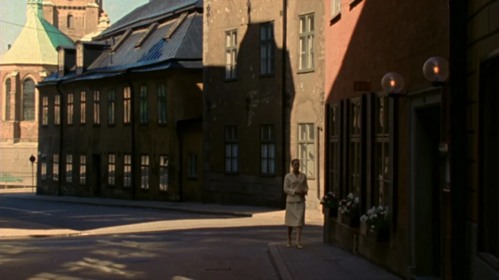 Face to Face (1976) movie still: wide — A historic European street corner features masonry buildings and a distant green-roofed spire. A woman in a…