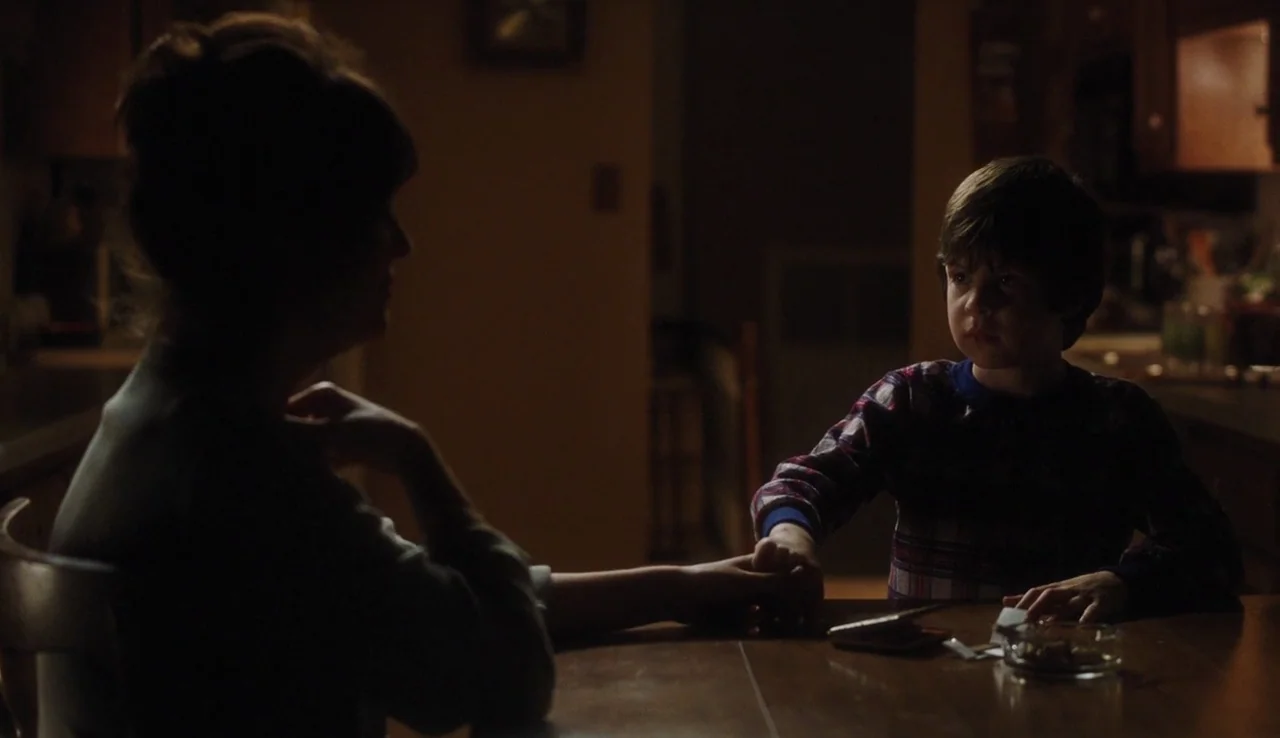 The Perks of Being A Wallflower (2012) movie still: medium — A dimly lit room features a wooden table where a woman and child sit. The boy holds the…