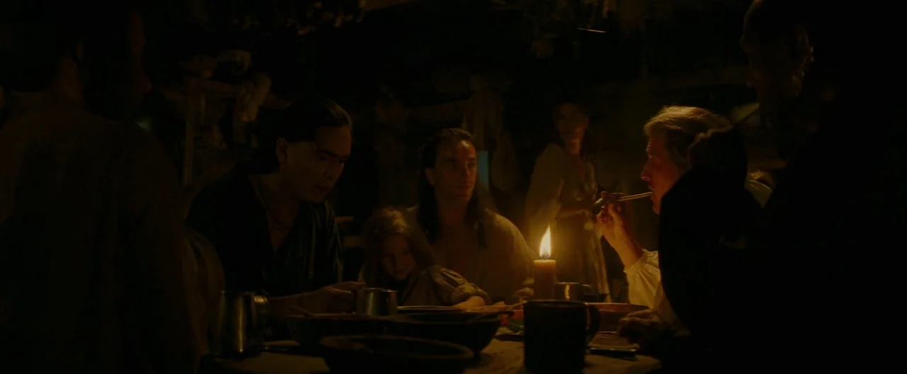 The Last of the Mohicans (1992) movie still: medium — A dark, rustic interior features multiple people gathered around a wooden table at night. A…