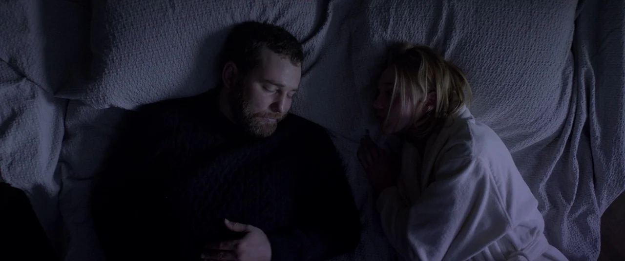 Loveless (2017) movie still: medium — A man and a woman lie on a textured grey blanket in dim light. The man is sleeping while the woman in a robe…