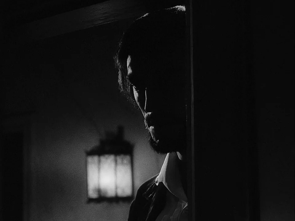 The Magician (1958) movie still: closeup — A dark hallway features a glowing lantern mounted on a distant wall. A man stands in the foreground,…