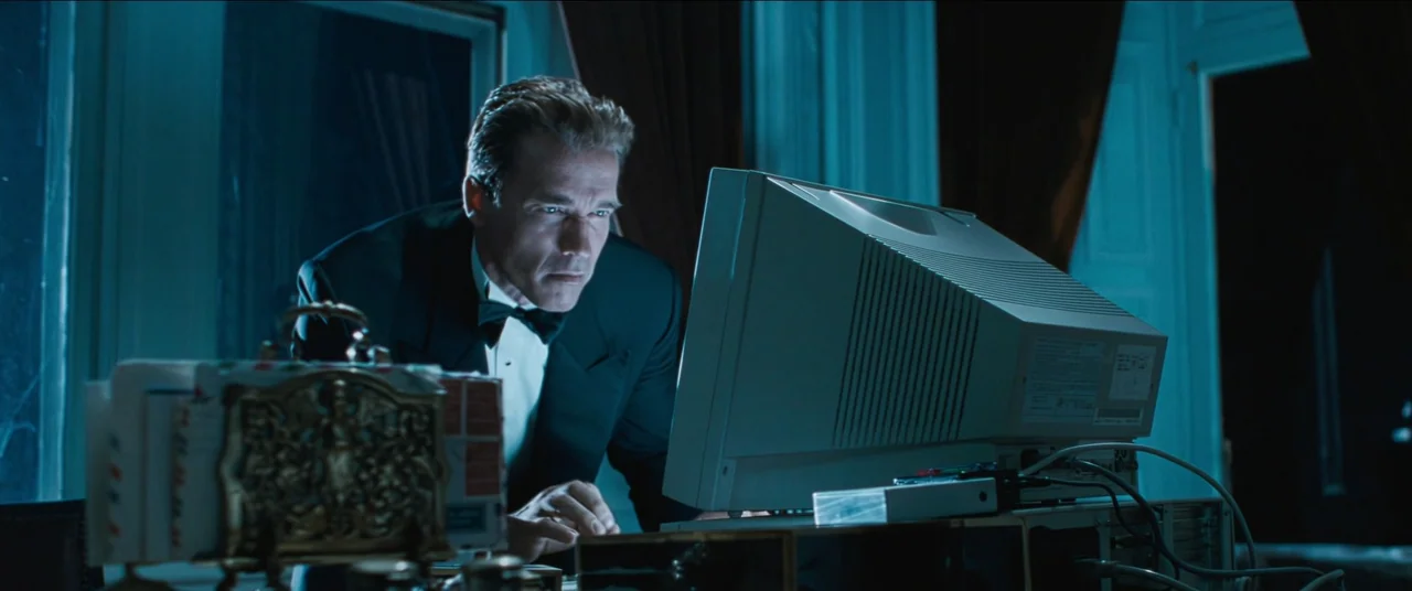 True Lies (1994) movie still: medium — A dimly lit room containing a desk with a bulky vintage computer monitor. A man in formal evening wear stares…