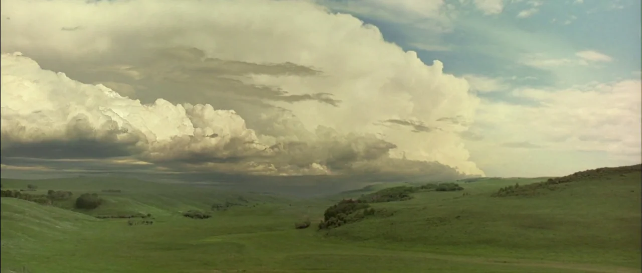 Open Range (2003) movie still: establishing — A vast expanse of green rolling hills extends under a massive, dense cloud formation. The sky is…