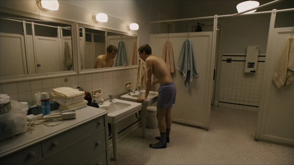 Youth In Revolt (2009) movie still: medium — A tiled restroom features two sinks, white stall doors, and hanging towels. A shirtless young man…