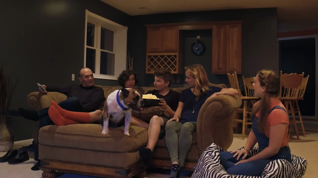 Sk8 Dawg (2018) movie still: medium — A living room interior features a large brown couch with four people and a dog. A group of individuals is…