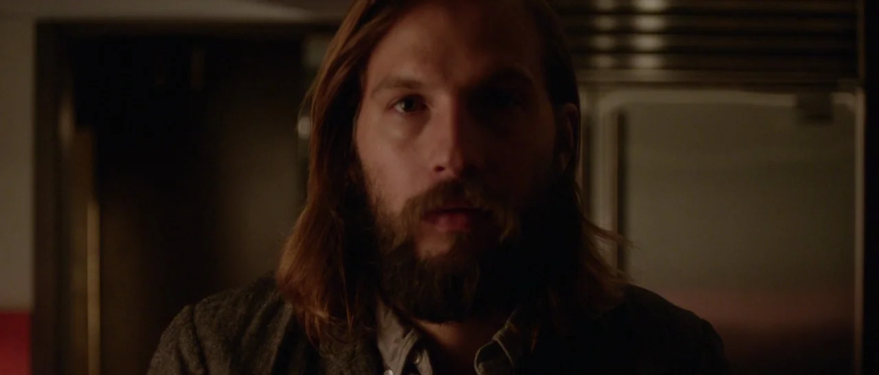 The Invitation (2015) movie still: closeup — A man stands in a dimly lit kitchen with stainless steel appliances in the back. The man looks forward…