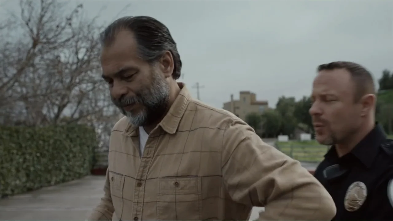 The Cleaner (2021) movie still: medium — A paved lot sits beneath an overcast sky with dry trees in the distance. An officer escorts a man in a tan…