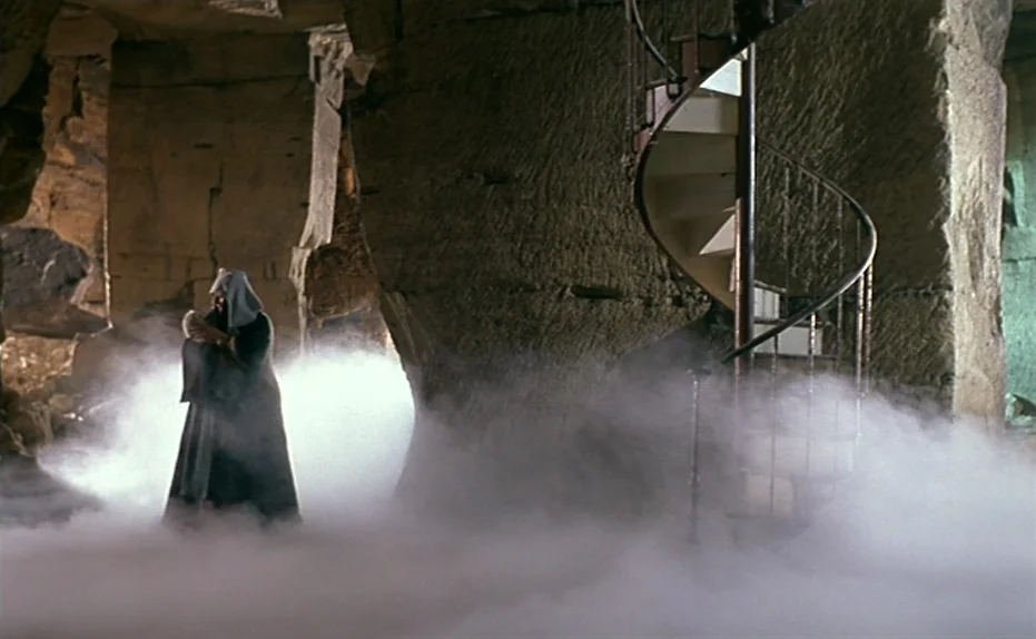 Life Is A Bed of Roses (1983) movie still: wide — A vast limestone cavern features rough-hewn stone pillars and a central metal spiral staircase. A…