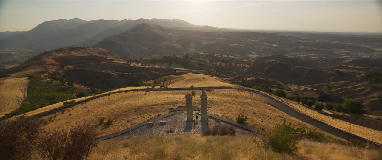 About Dry Grasses (2023) movie still: establishing — A wide, elevated view looks across a dry, hilly landscape towards distant hazy mountains. Two…